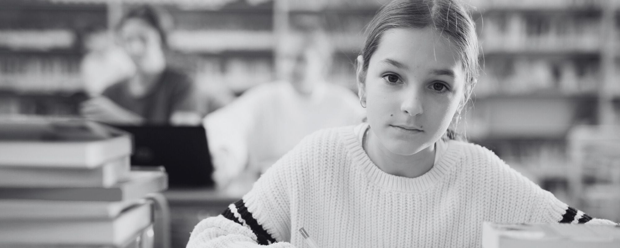 Black and white photo of a Middle school student, girl, in sitting in library, staring directly at camera, serious, banner image 2000x800